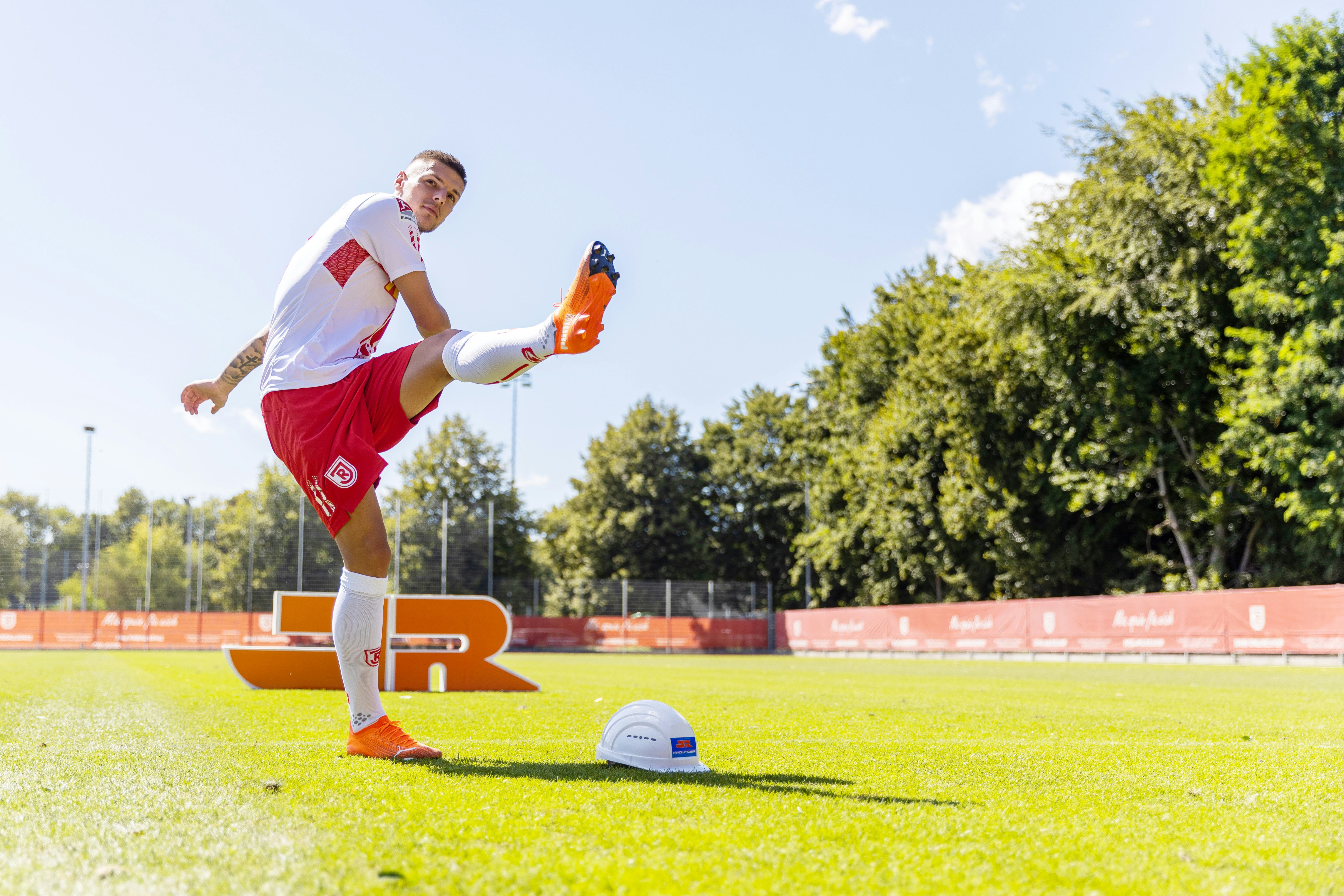 Spieler des SSV Jahn Regensburg auf Fußballplatz, Rädlinger Bauhelm auf dem Rasen, im Hintergrund Rädlinger Logo