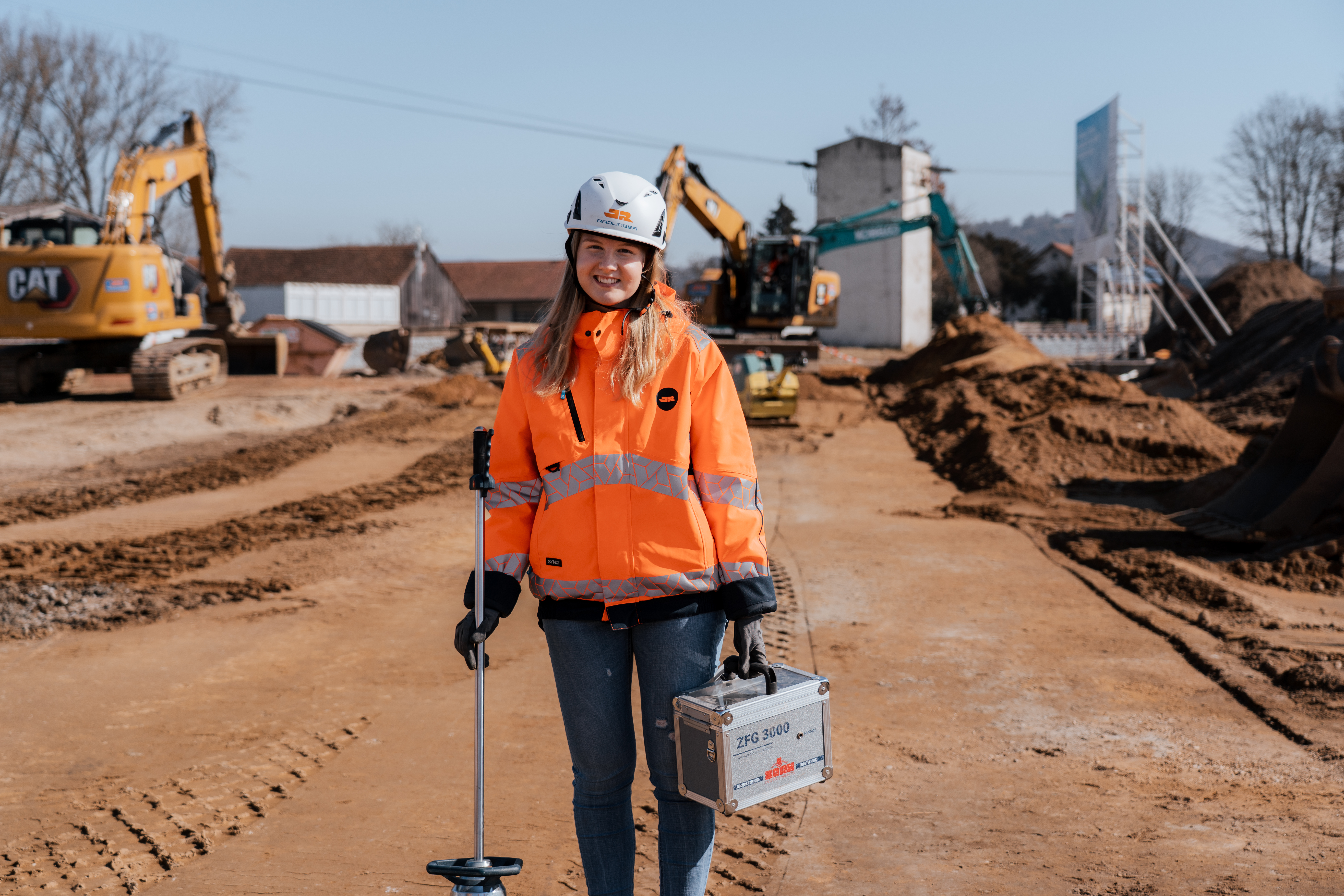 Eine Auszubildende zur Baustoffprüferin auf einer Erdbau-Baustelle, im Hintergrund Bagger