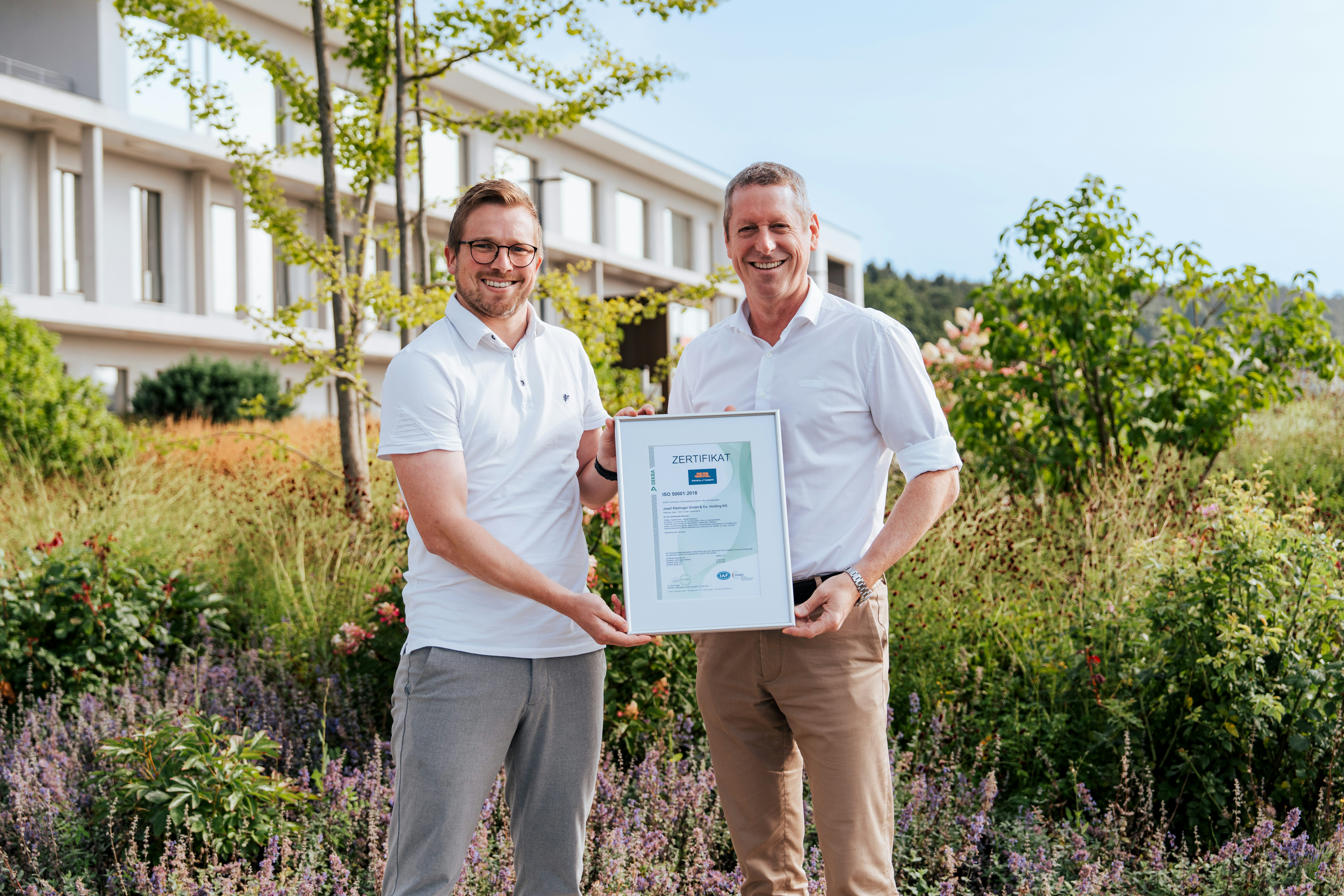 Martin Wagner (links), Energiemanagement-Beauftragter bei der Josef Rädlinger Unternehmensgruppe, und Geschäftsführer Rüdiger Altmann präsentieren das ISO 50001-Zertifikat für das Energiemanagementsystem vor dem modernen Bürogebäude der Hauptverwaltung in Cham – ein wichtiger Meilenstein für Nachhaltigkeit und Energieeffizienz.
