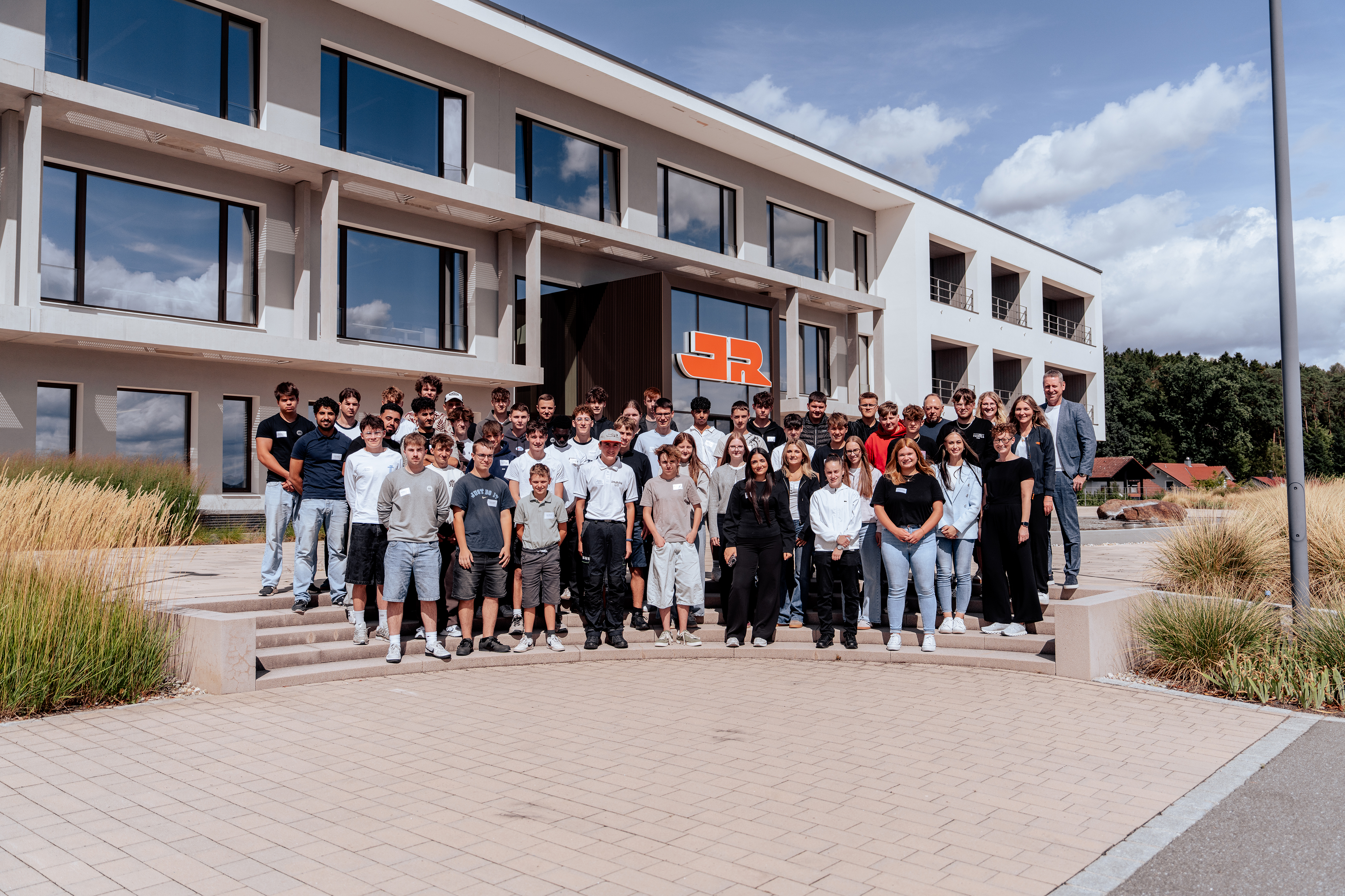 Gruppenfoto vor dem Verwaltungsgebäude der Josef Rädlinger Unternehmensgruppe: Zahlreiche Auszubildende und Mitarbeitende stehen vor dem Haupteingang mit JR-Logo auf den Treppenstufen der Unternehmenszentrale.