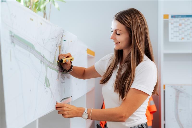 Frau steht an einer Wand mit großformatigen technischen Plänen und markiert Linien mit einem Stift