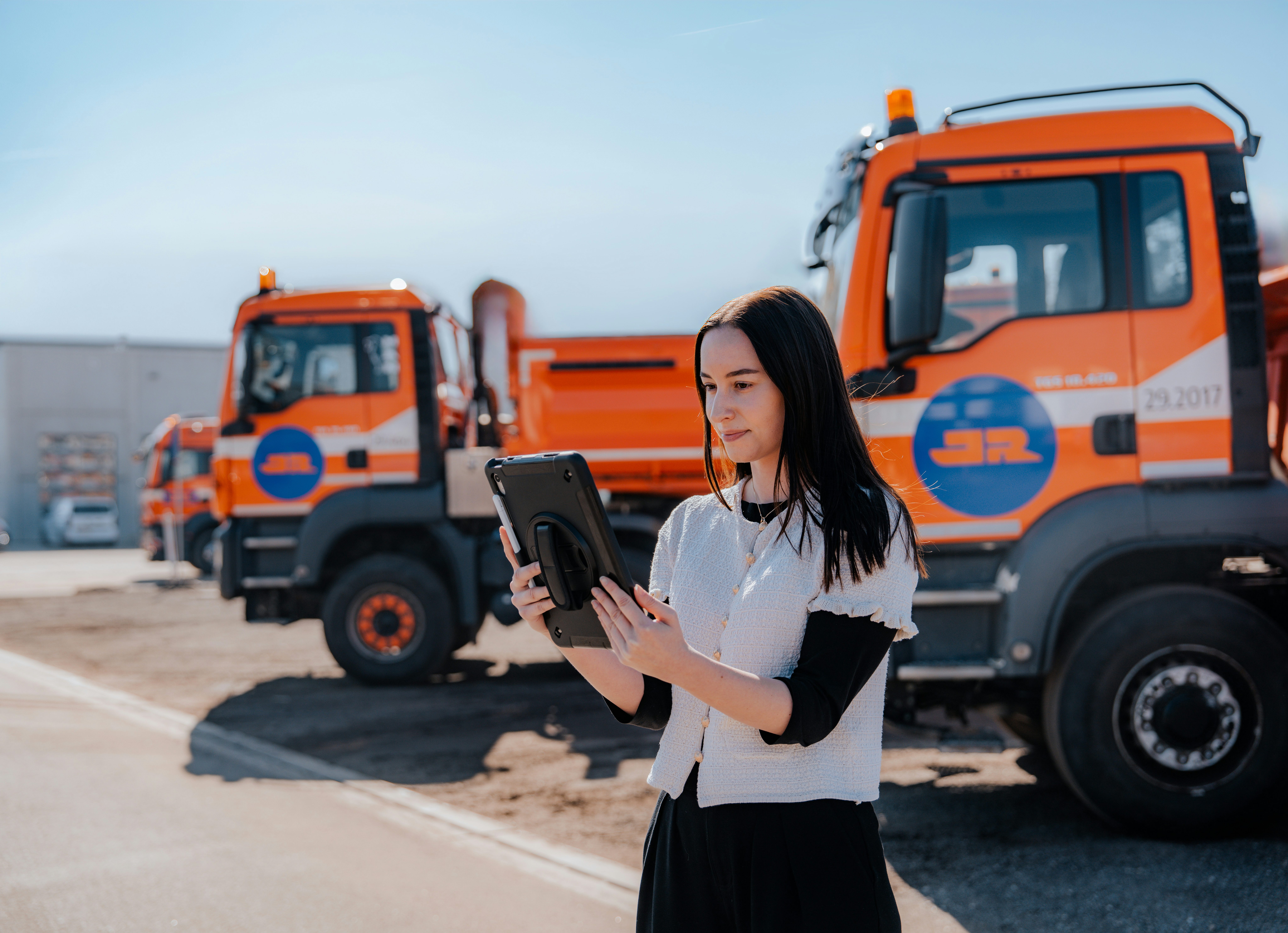 Person hält ein Tablet vor mehreren orangefarbenen Lkw und arbeitet im Außenbereich an der digitalen Disposition