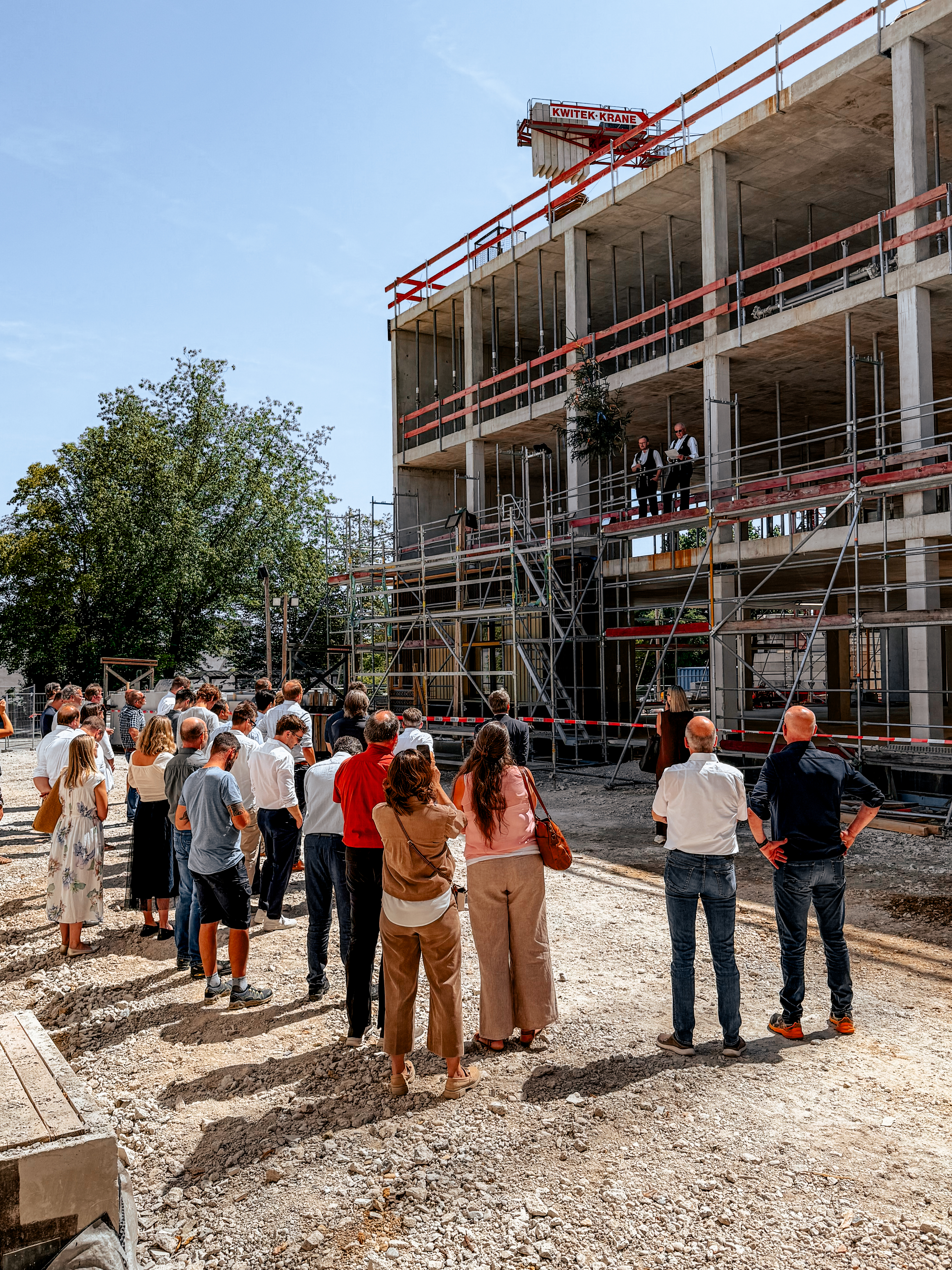 Die Projektbeteiligten stehen vor dem Rohbau, auf dem Gerüst wird der Richtspruch verlesen.