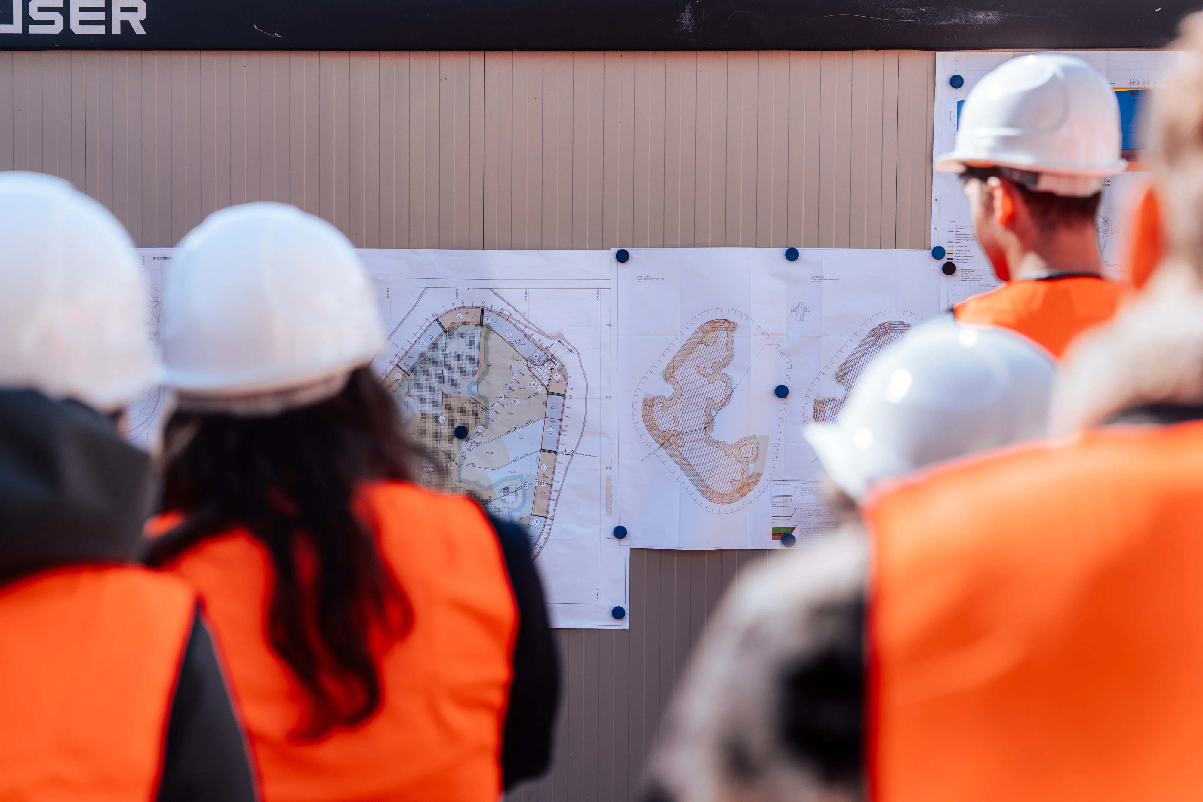 Mehrere Personen mit Schutzhelmen und Warnwesten betrachten aus kurzer Entfernung technische Lagepläne, die an einer Informationstafel auf einer Baustelle befestigt sind.