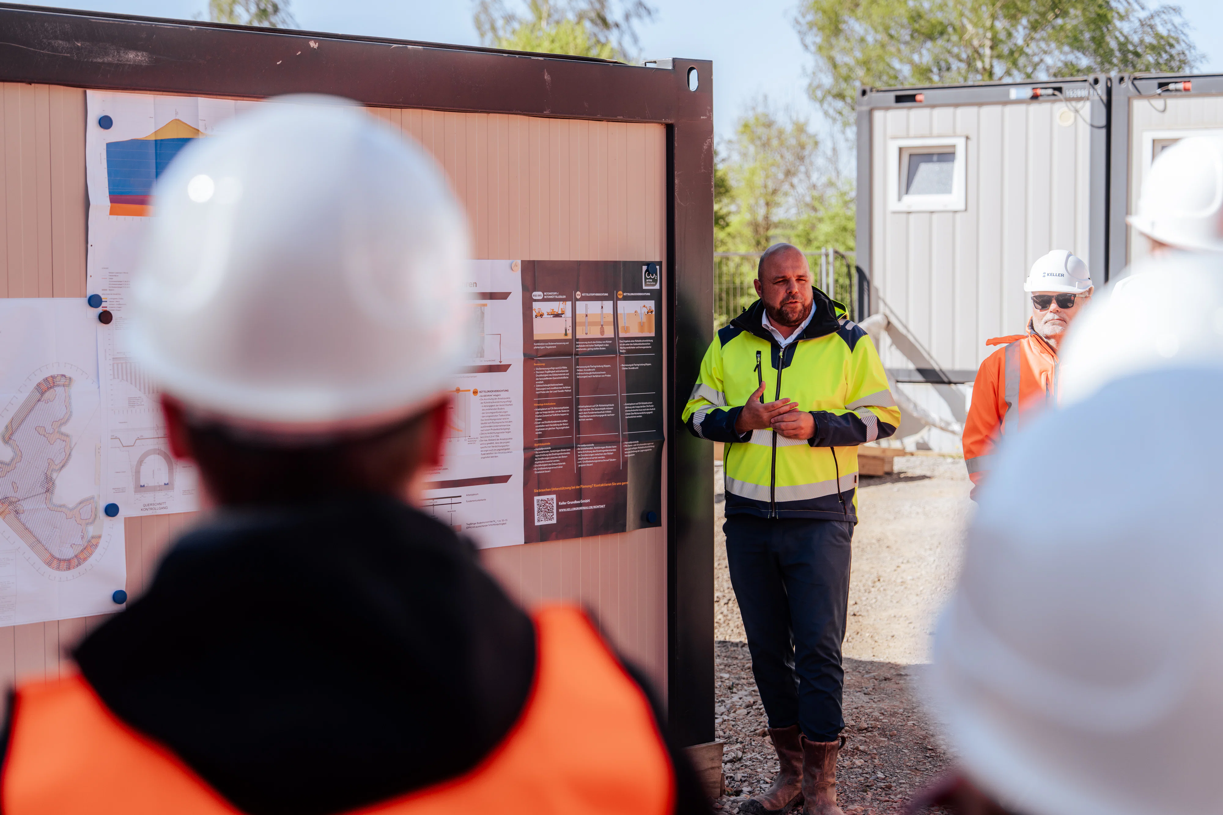 Person in Warnschutzkleidung erläutert einer Gruppe von Besuchenden mit Schutzhelmen Informationen an einer Baustellen-Infotafel mit Plänen und Grafiken. Im Hintergrund sind Baustellencontainer zu sehen.
