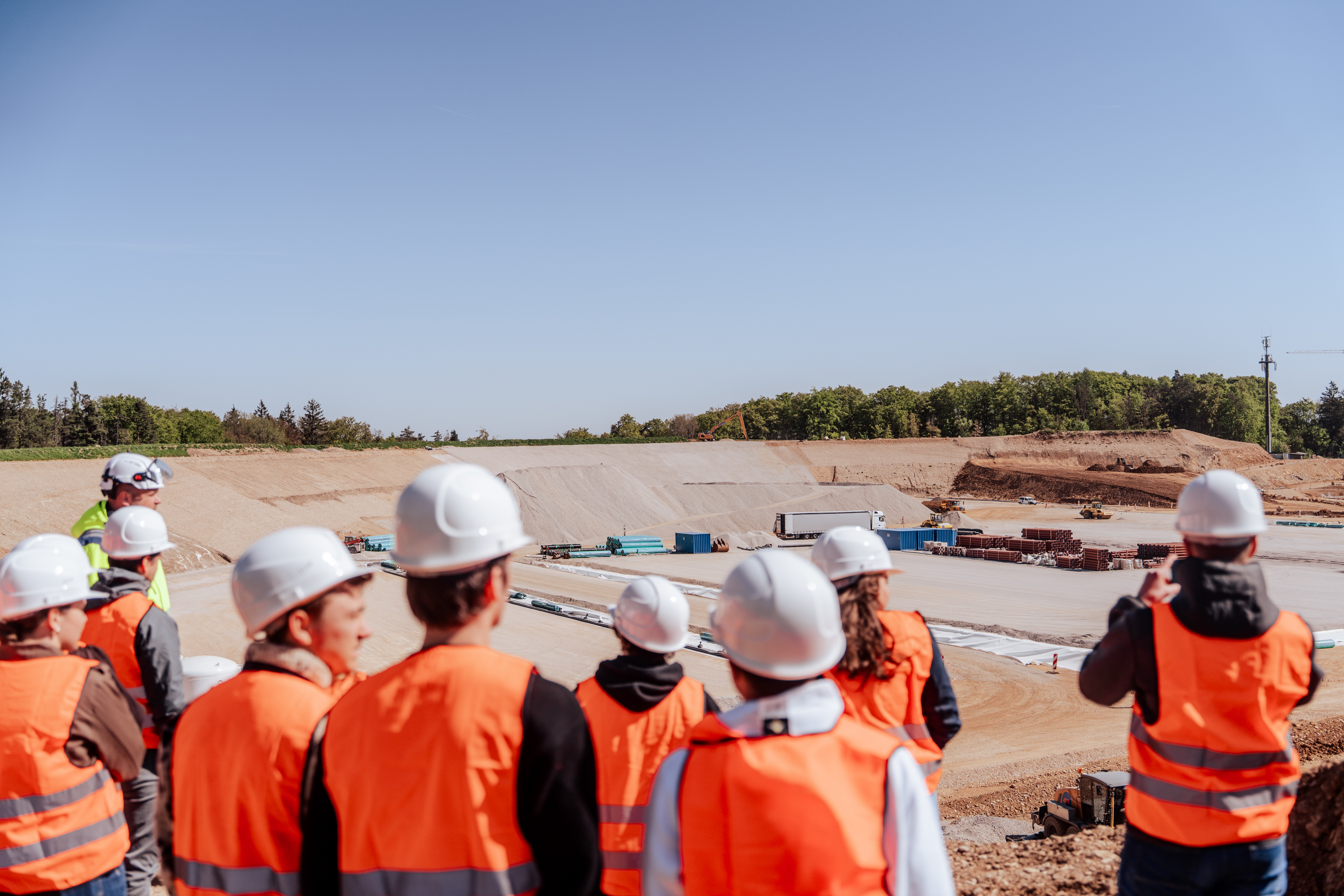 Gruppe von Personen mit Schutzhelmen und Warnwesten blickt auf eine großflächige Baustelle mit Erdbauarbeiten, Lagerflächen und Baumaschinen. Im Hintergrund sind Böschungen und ein bewaldeter Bereich zu sehen.