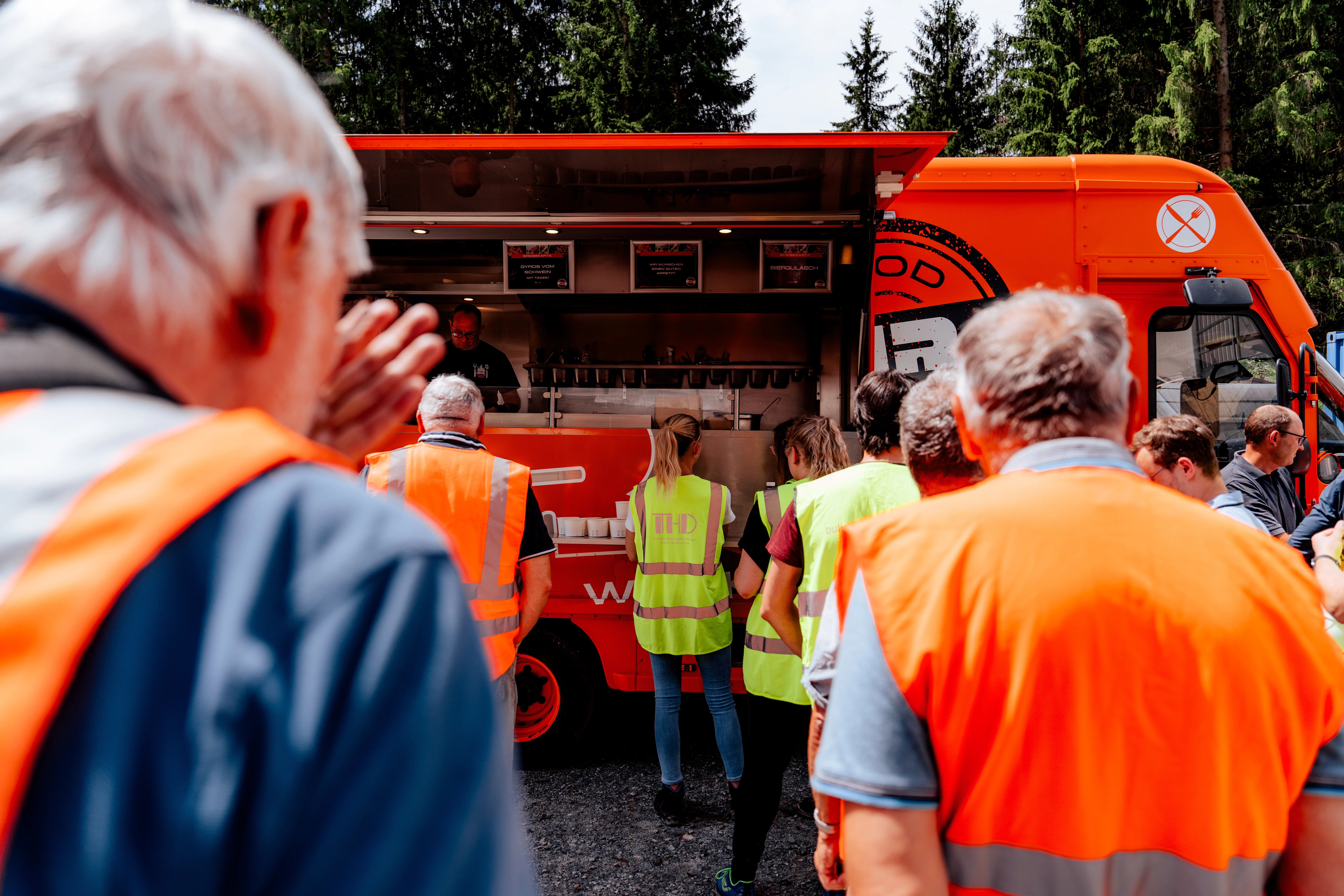 Mehrere Personen in Warnwesten stehen vor einem orangefarbenen Foodtruck im Freien und warten auf Verpflegung. Im Hintergrund sind Bäume und der geöffnete Ausgabebereich des Fahrzeugs zu sehen