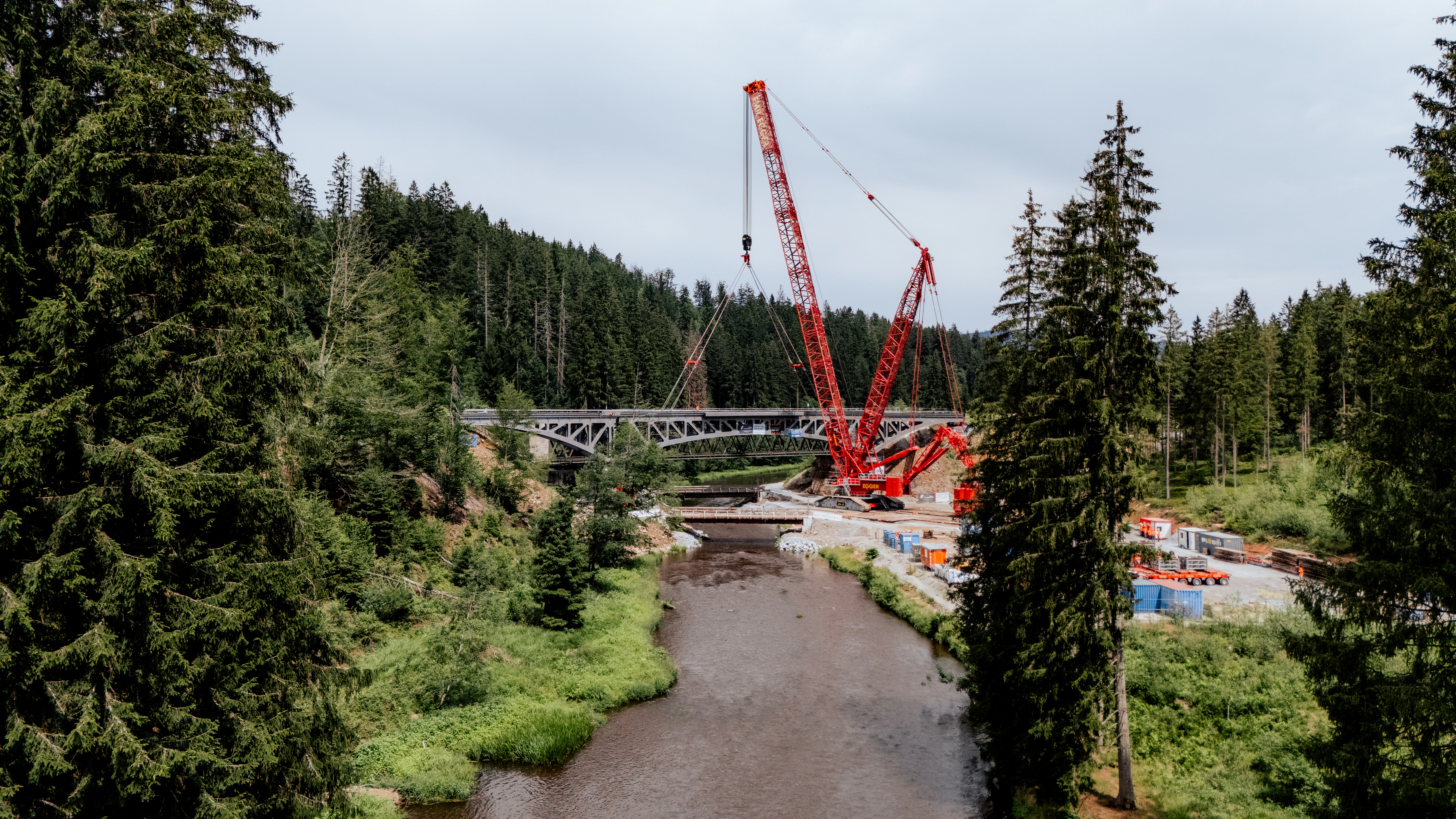 Luftaufnahme einer Brückenbaustelle in bewaldeter Landschaft: Ein großer roter Raupenkran hebt ein Brückenelement über einem Fluss ein, während Fahrzeuge, Baumaschinen und Baustelleneinrichtungen im Talbereich angeordnet sind.