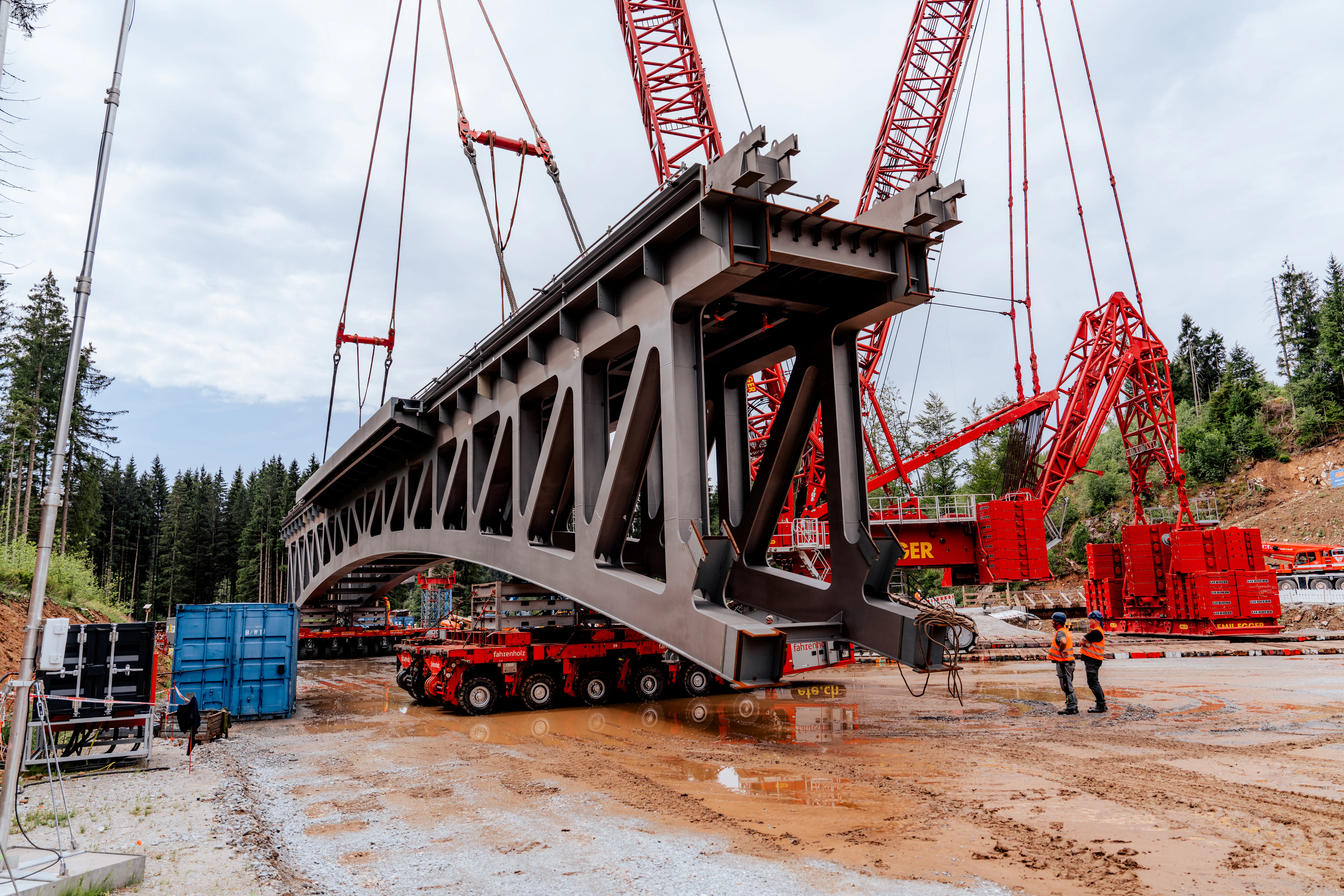 Großes Stahlbrückenelement wird auf einer Baustelle mit mehreren roten Raupenkränen angehoben. Das Brückensegment schwebt über einem Schwerlasttransporter, während Mitarbeitende den Einhub begleiten.