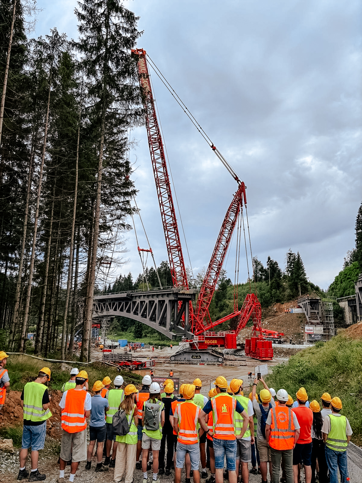 Mehrere Personen mit Schutzhelmen und Warnwesten beobachten den Einhub eines Stahlbrückenelements. Ein großer roter Raupenkran hebt die Brücke auf einer Baustelle inmitten eines bewaldeten Tals an.
