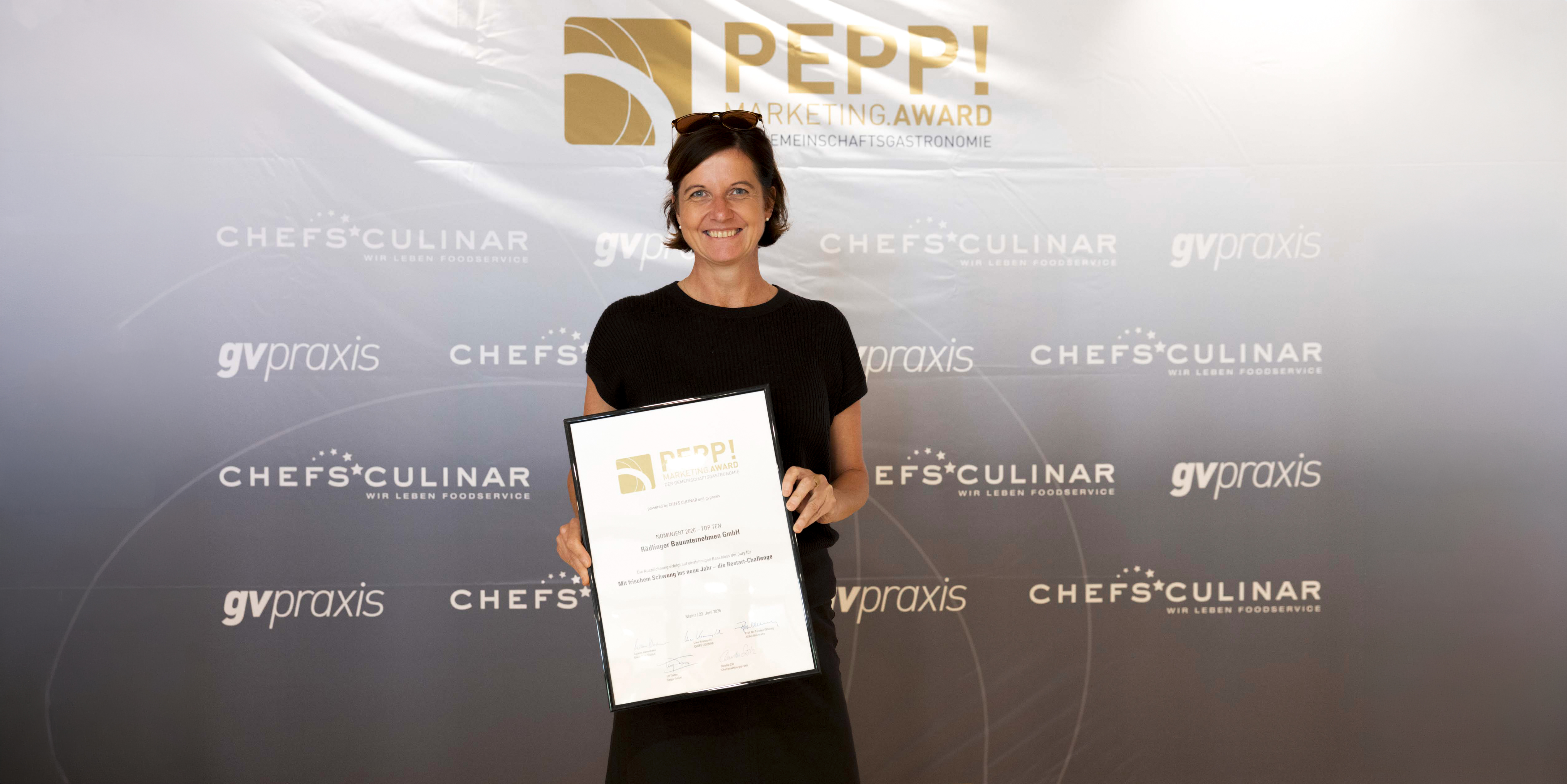 Person steht vor einer Event‑Fotowand mit Logos von ‚PEPP! Marketing Award‘, ‚CHEFS CULINAR‘ und ‚gvpraxis‘ und hält eine gerahmte Urkunde in den Händen.