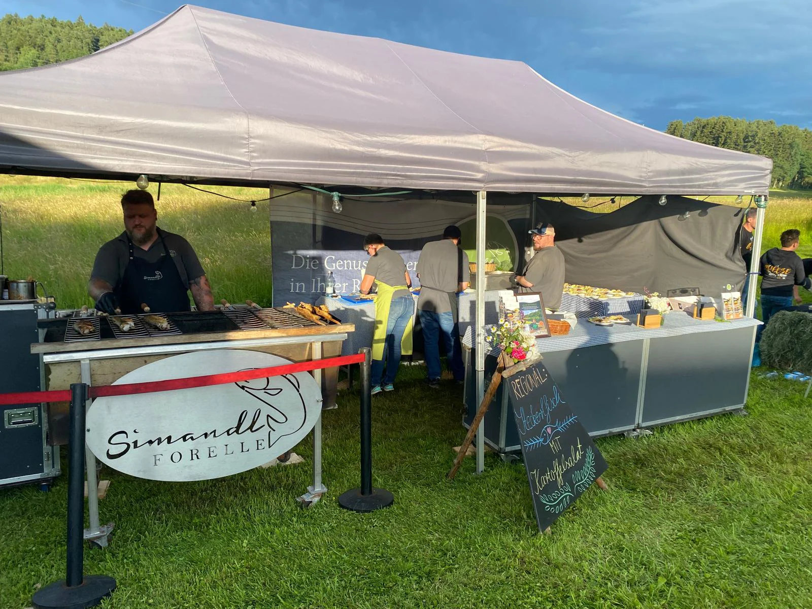 Aufnahme eines Verkaufszeltes mit mehreren Menschen und einem Grill, auf dem Steckerlfische gegrillt werden