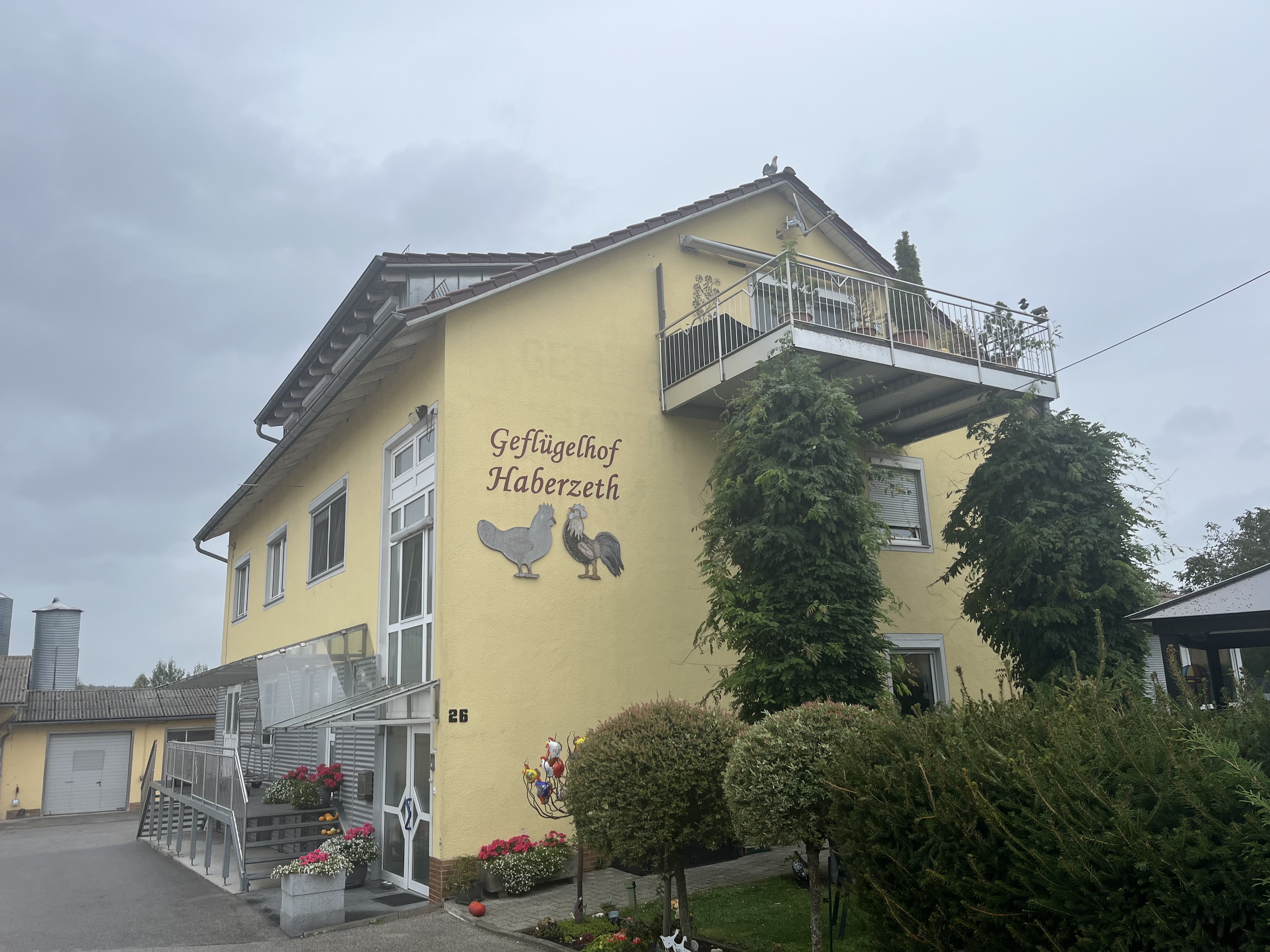Gelb gestrichenes Wohn- und Betriebsgebäude des Geflügelhofs Haberzeth mit Balkon, begrünten Fassadenelementen und bepflanztem Eingangsbereich. An der Fassade ist die Aufschrift ‚Geflügelhof Haberzeth‘ mit Huhn- und Hahnmotiv angebracht.