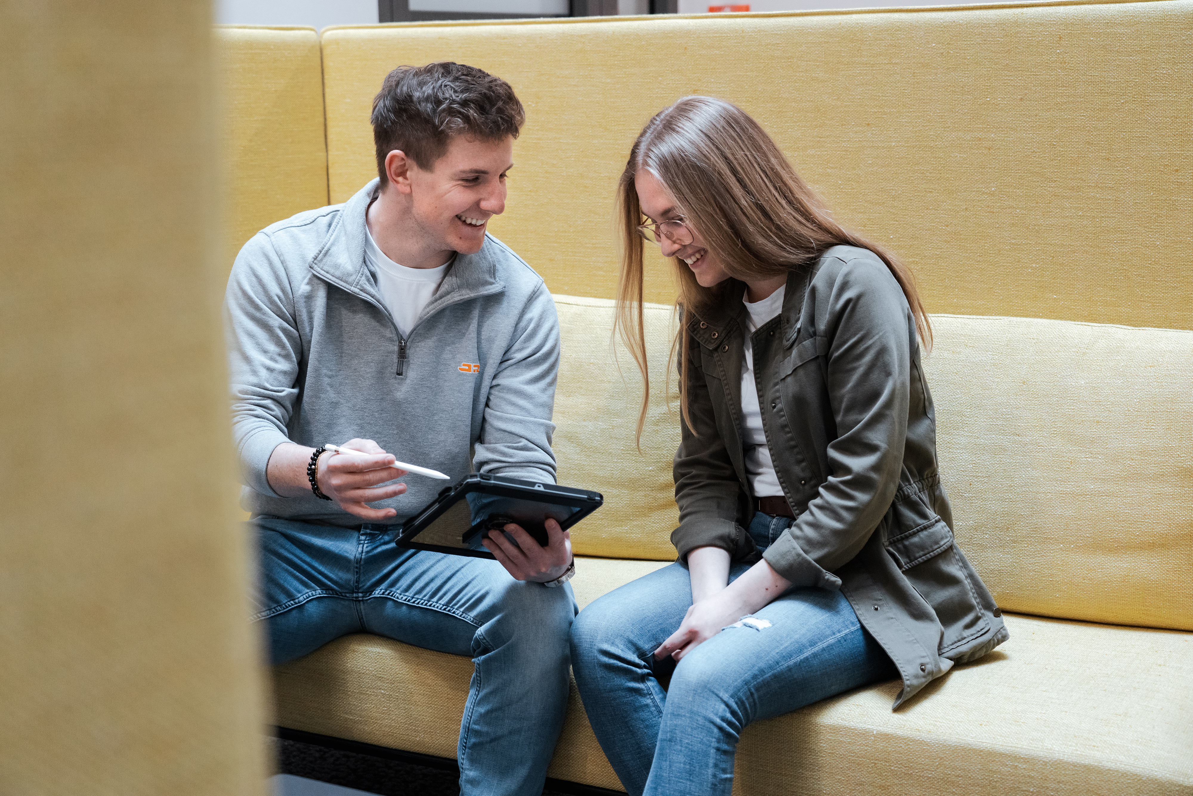 Rädlinger Mitarbeiter und Mitarbeiterin sitzen auf einer Couch und unterhalten sich, Mitarbeiter zeigt Mitarbeiterin etwas auf dem Tablet