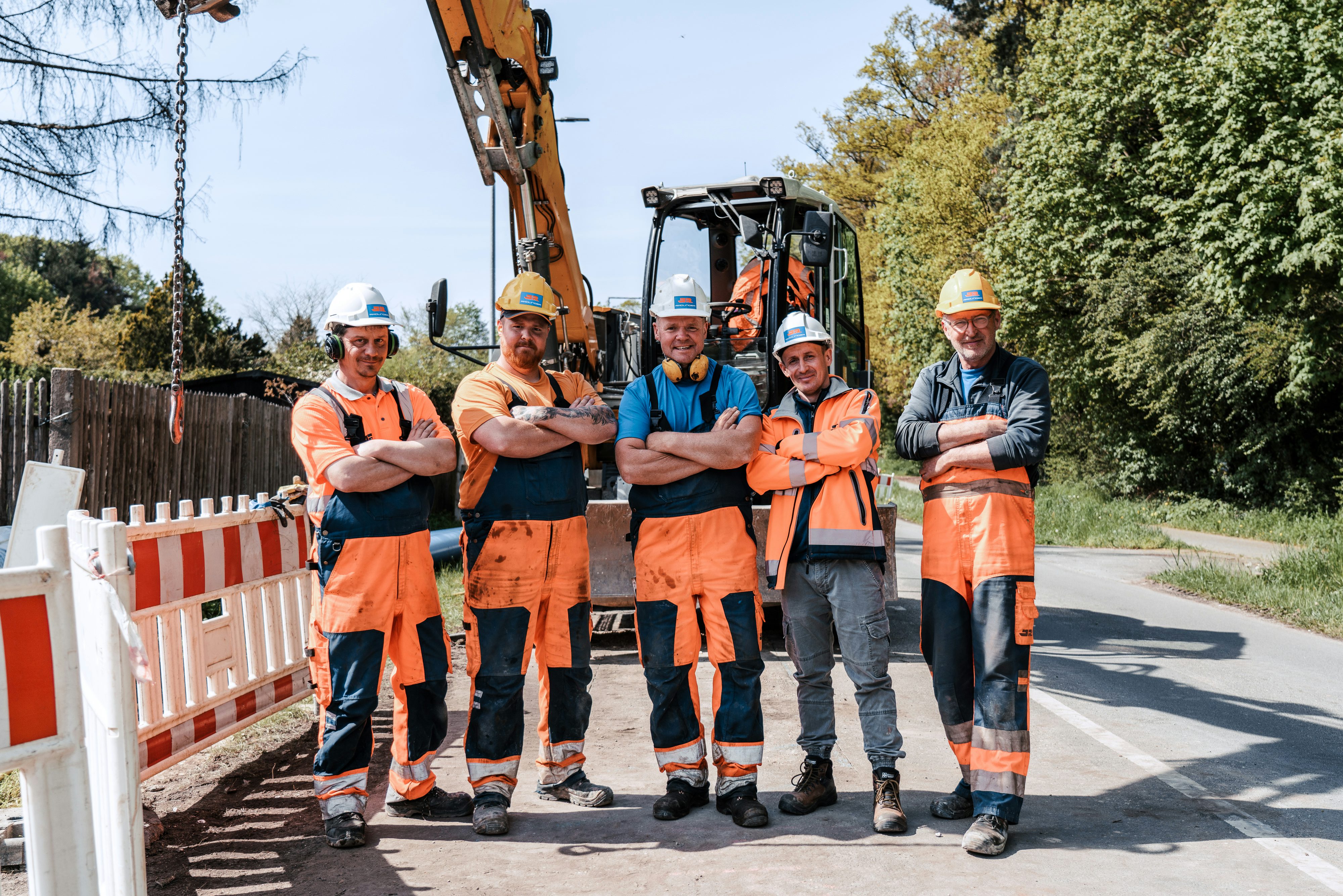 Fünf Baustellenarbeiter lächelnd und mit verschränkten Armen auf der Baustelle vor Bagger