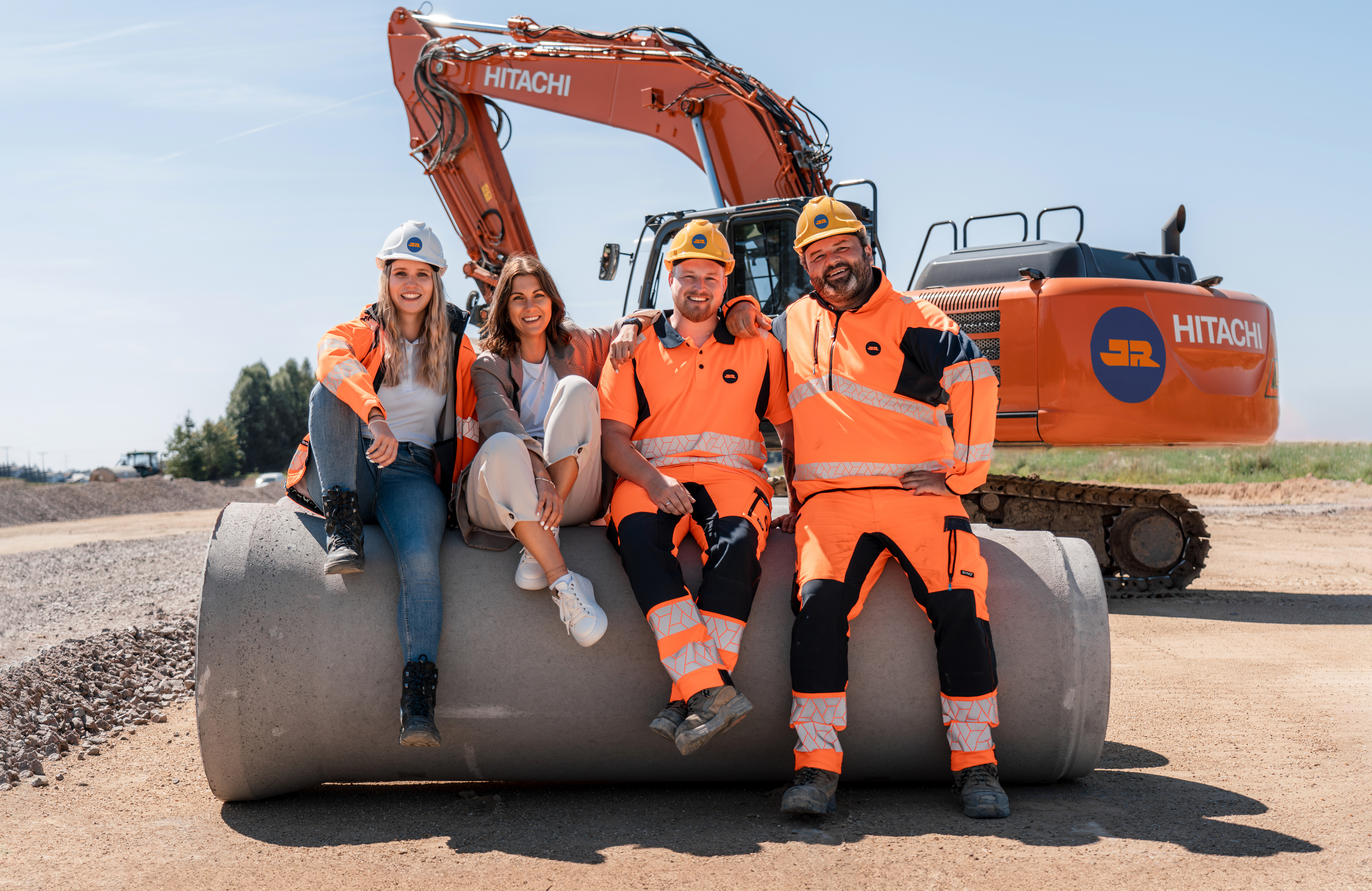 Vier JR Mitarbeitende sitzen nebeneinander auf einem Betonrohr auf der Baustelle, im Hintergrund ein Bagger