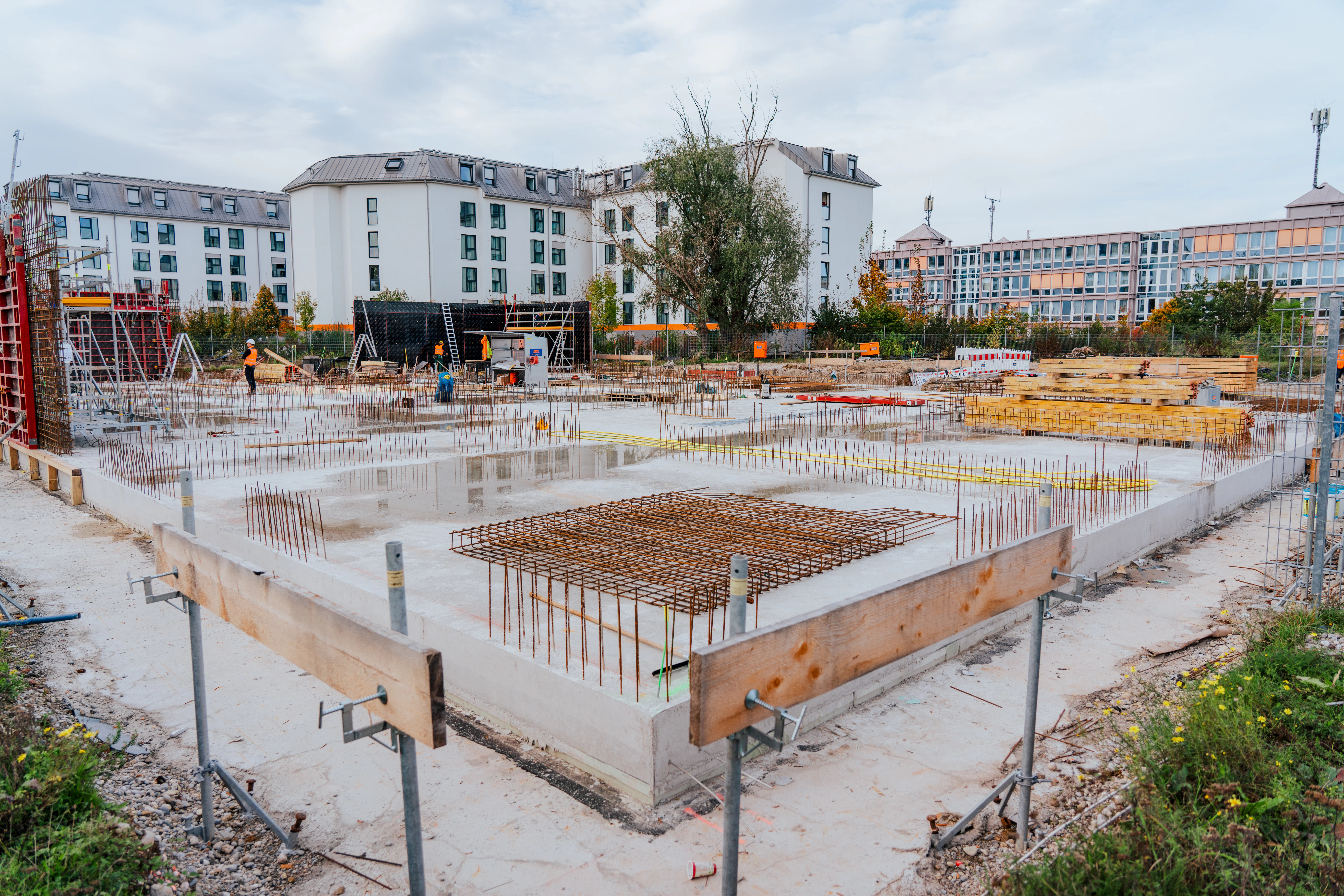 Bewehrungsstahl und Holz auf dem Betonfundament für das Gebäude. Im Hintergrund einige JR Mitarbeitende sowie Schalungselemente