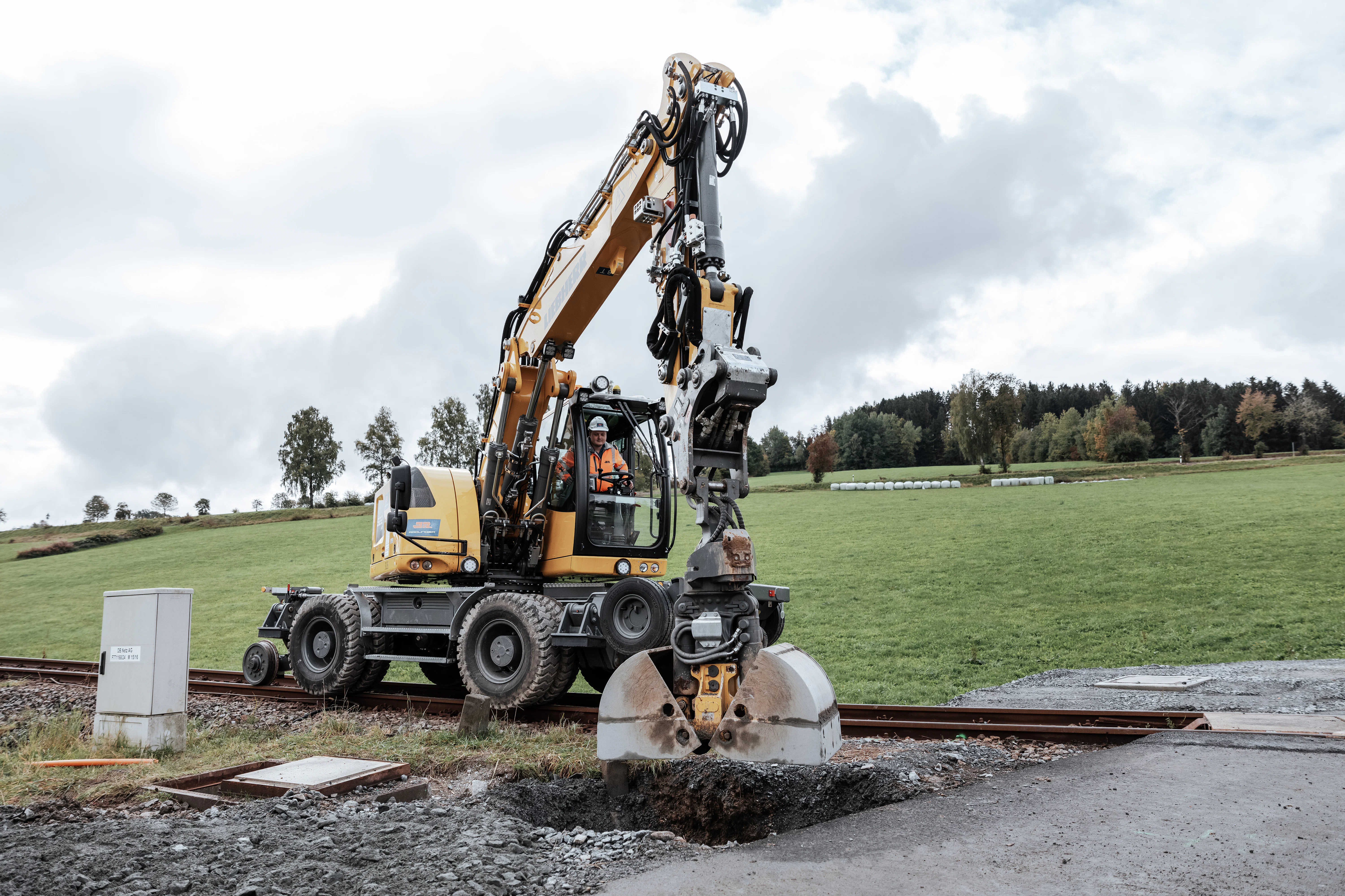 Zweiwegebagger steht auf dem Gleis und baggert Grube an einem Bahnübergang aus