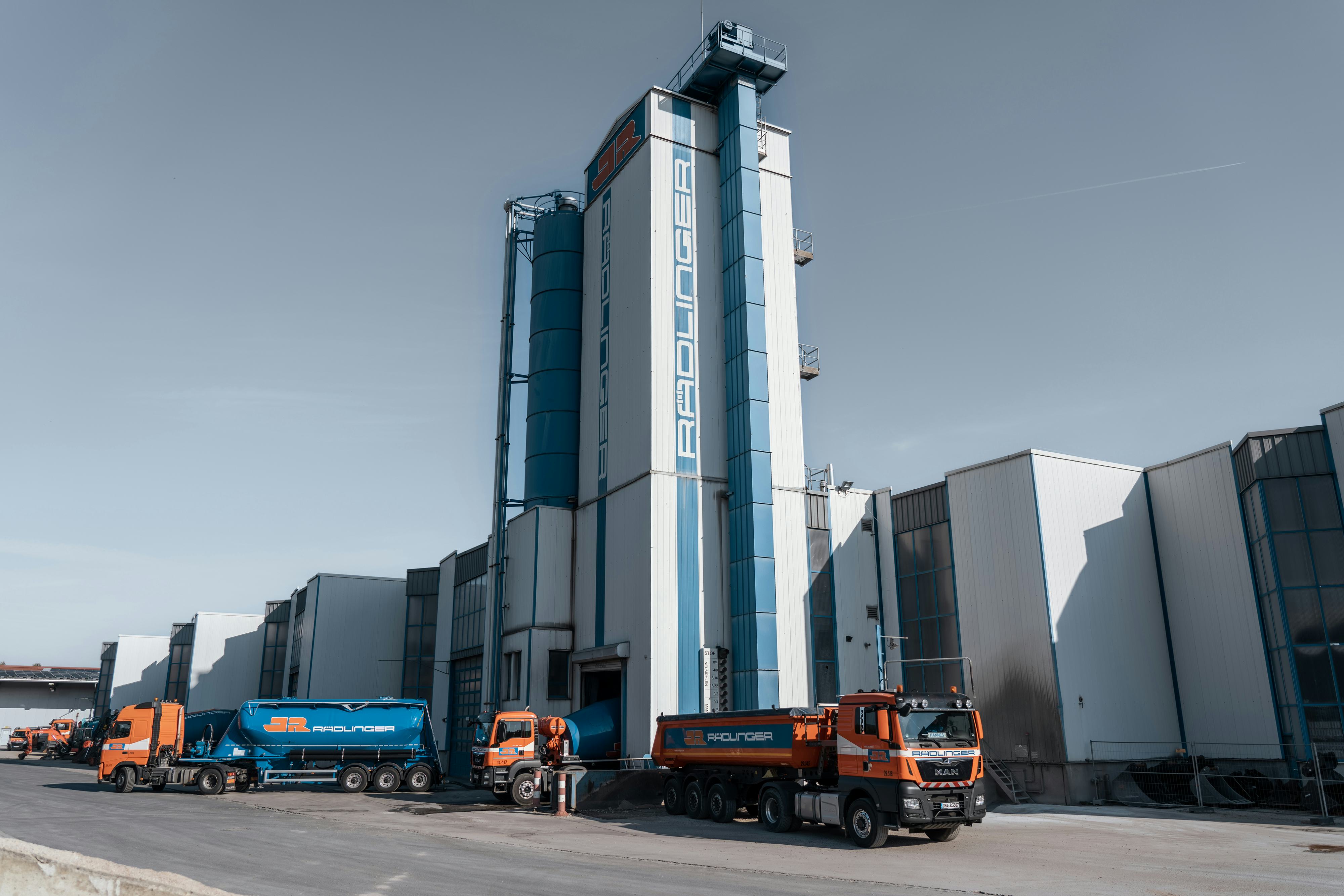 Rädlinger Standort in Weiding: Turmanlage des Betontransportwerks von außen, davor verschiedene Maschinen wie Betonmischer, Sattelzug und Lkw mit Kipper